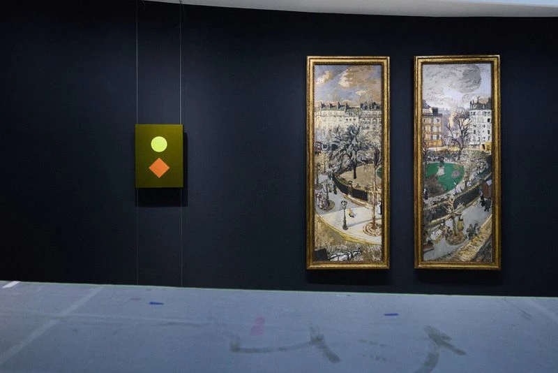 Indoor shot of framed art pieces on a dark wall, including cityscapes and abstract geometric art, displayed in an art gallery.
