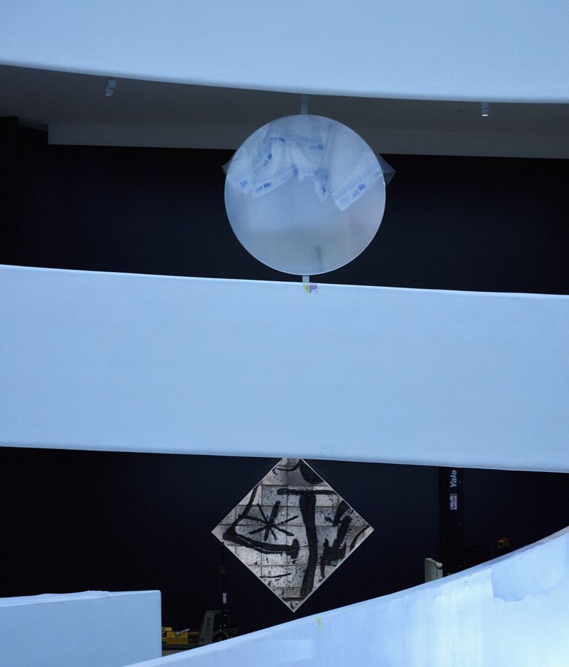 A translucent balloon-like sculpture hangs between white balcony railings in a museum space with black walls, creating a modern art display.
