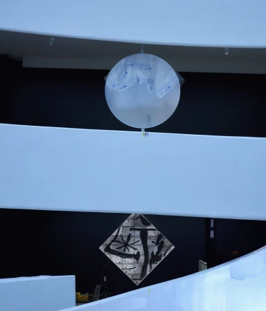 A translucent balloon-like sculpture hangs between white balcony railings in a museum space with black walls, creating a modern art display.