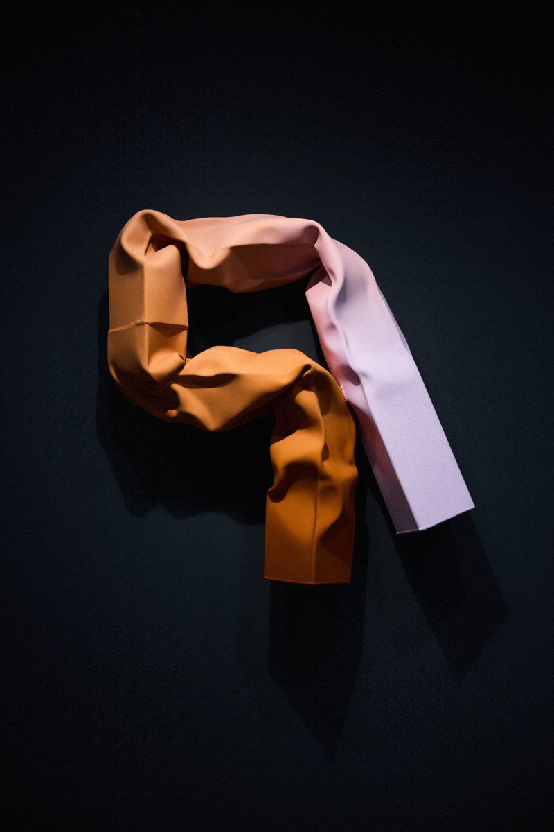 Abstract photograph of a draped, two-toned fabric with orange and lilac sections against a dark gray background in a studio setting.