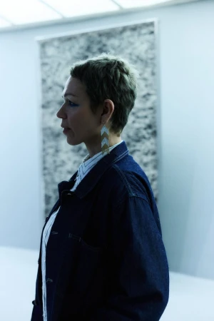 Profile view of a woman in a denim jacket and long earrings standing indoors in front of a gray and white abstract background art piece.