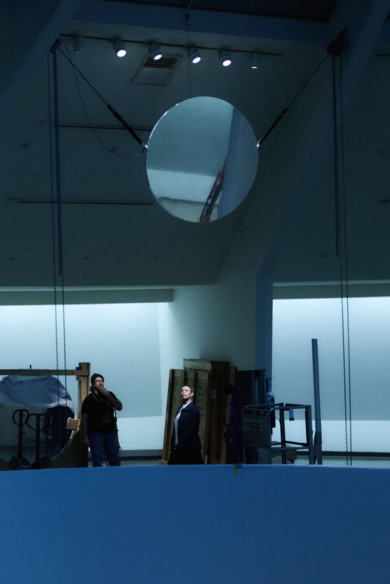 A large, suspended disc reflects the interior of a spacious hall with two men standing near some wood palettes. Indoor lighting.