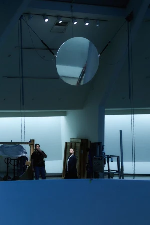 A large, suspended disc reflects the interior of a spacious hall with two men standing near some wood palettes. Indoor lighting.