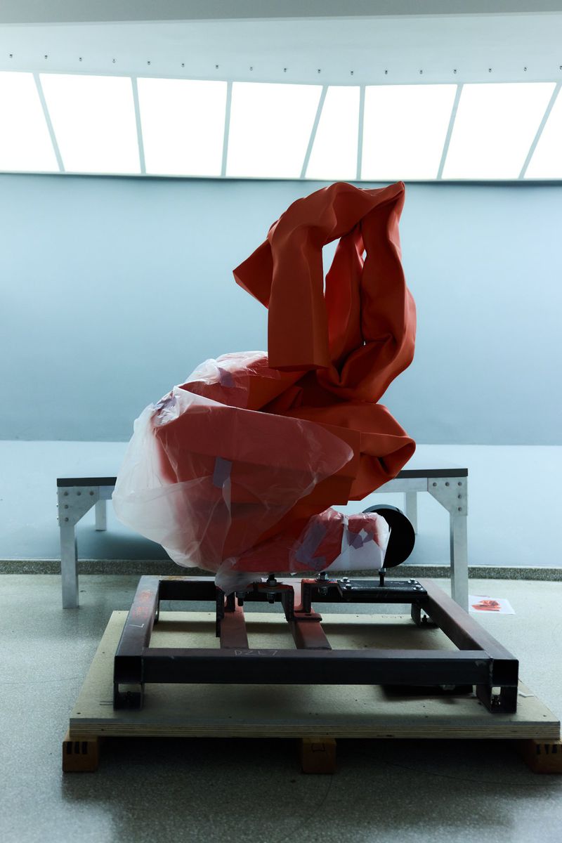 Abstract orange fabric sculpture on a wooden pallet in a bright studio. Modern art installation with industrial elements and white background.
