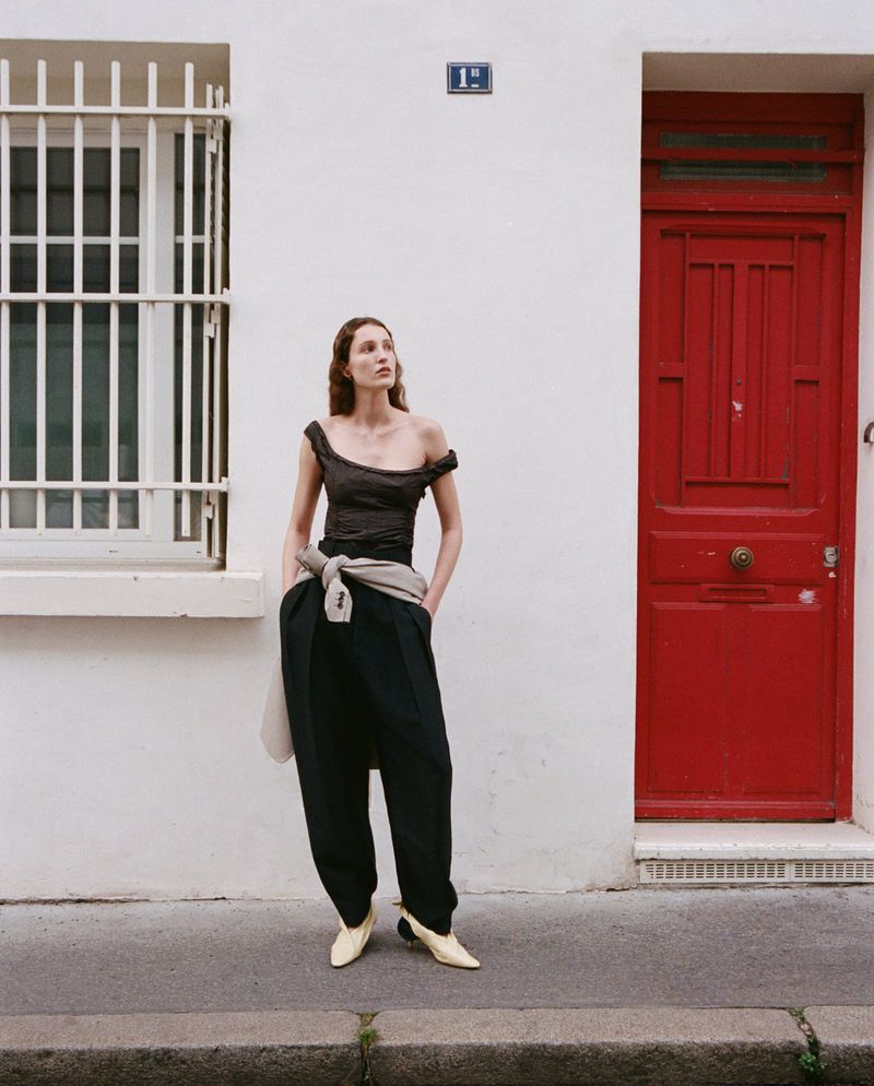 Adult woman poses in off-shoulder top and black pants near white building with red door in editorial fashion photograph outdoors.