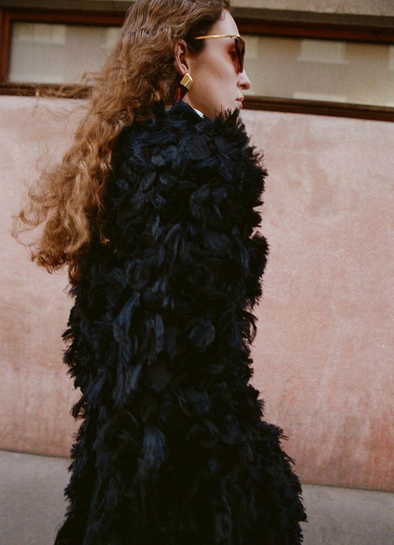 Stylish woman in a feathered coat walks outdoors with sunglasses and statement earrings, capturing a fashion-forward editorial look.