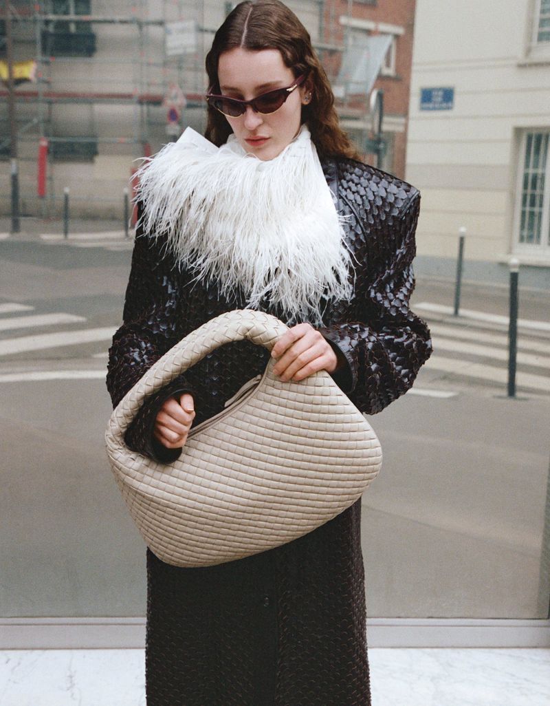 A woman in sunglasses and a textured brown coat stands on a city street holding a beige, woven handbag.