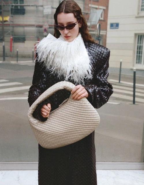 A woman in sunglasses and a textured brown coat stands on a city street holding a beige, woven handbag.