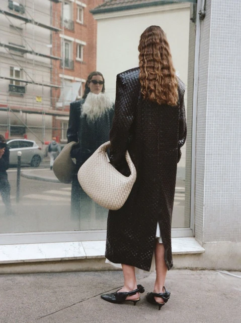 Fashionable adult woman in a long dark coat with a woven bag, seen in a mirror reflection in an urban setting on a city street.