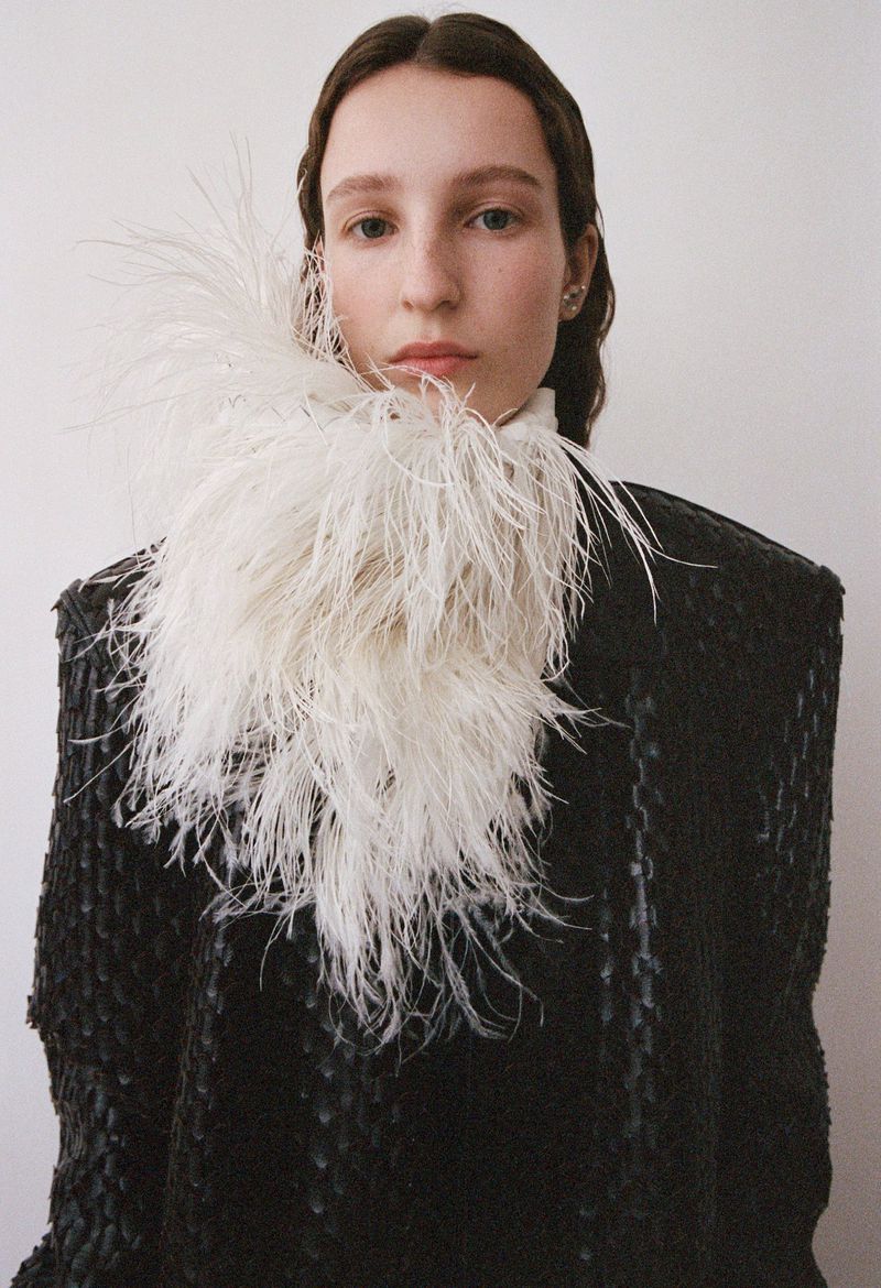 Fashion portrait of a young woman with light skin, wearing a black coat with a white feathered collar against a white background.