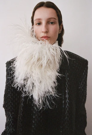 Fashion portrait of a young woman with light skin, wearing a black coat with a white feathered collar against a white background.