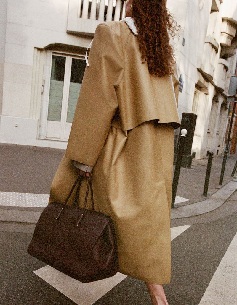Woman in a trench coat walking on a crosswalk holding a brown tote bag, with buildings and street lamps in the background.