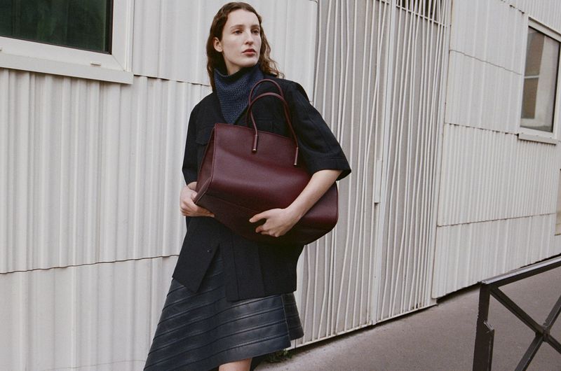 A fashionable woman in a black jacket and grey skirt holds a burgundy tote bag outdoors against a background of white metal siding.