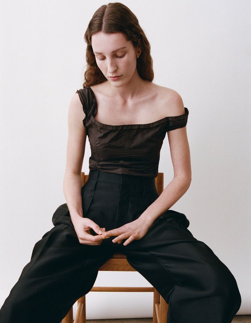 A stylish young woman with long brown hair poses in a studio wearing a dark top and high-waisted black pants while seated on a wooden chair.