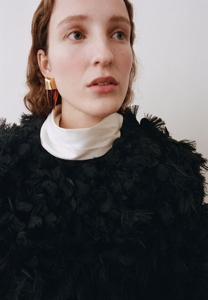 Close-up of a fair-skinned young woman with auburn hair wearing gold earrings and a dark, textured garment in a light studio setting.
