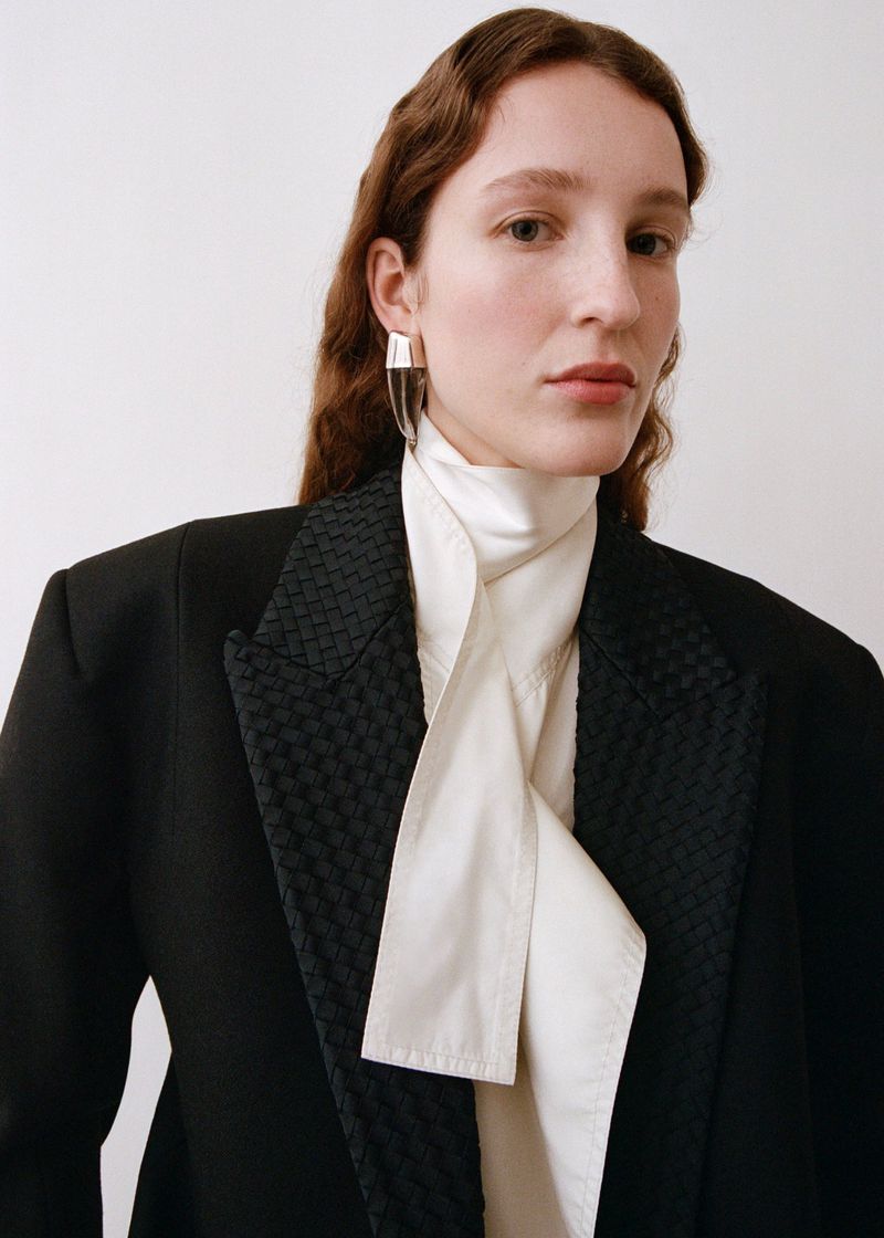 Portrait of an adult woman with wavy light-brown hair, wearing a black blazer and a silk blouse, set against a white backdrop.