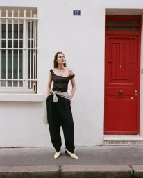 Model standing outdoors wearing a dark top, black trousers, and light boots against a white wall with a red door.