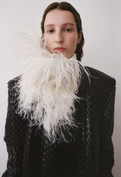 Model wearing a textured black jacket with a large white feather trim around the neck against a white wall.