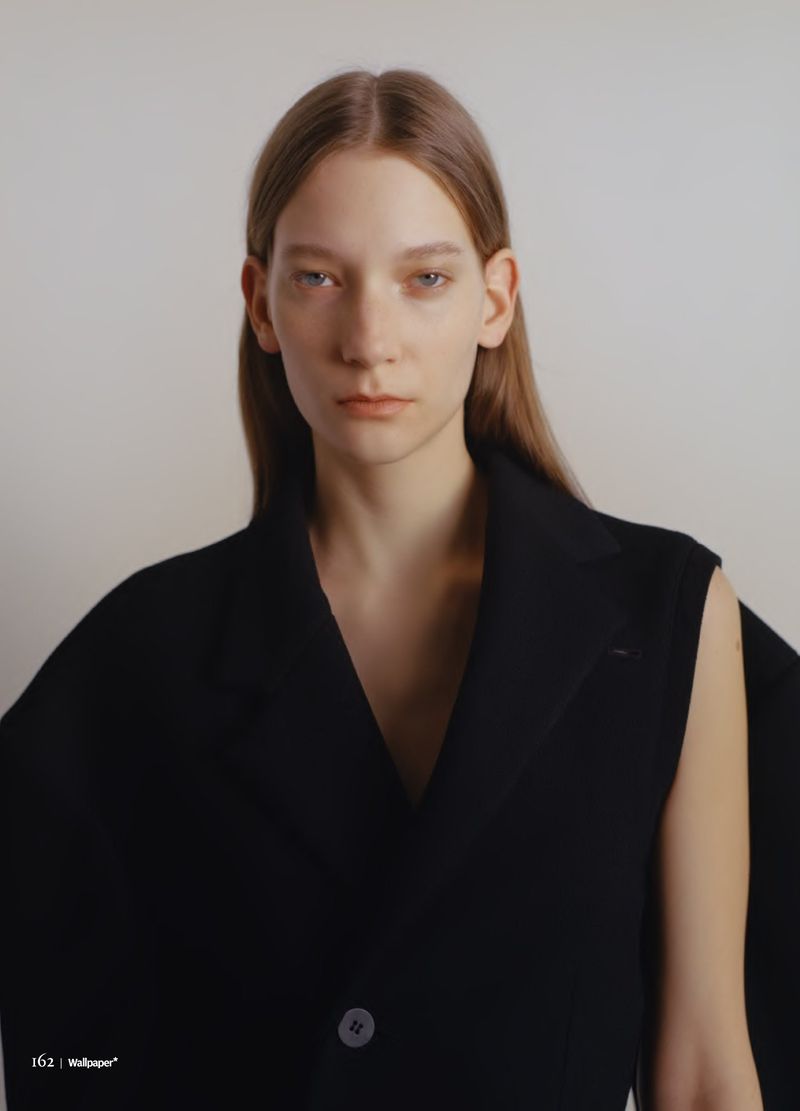 Close-up portrait of a young adult woman with long brown hair wearing a black sleeveless blazer against a neutral off-white backdrop.