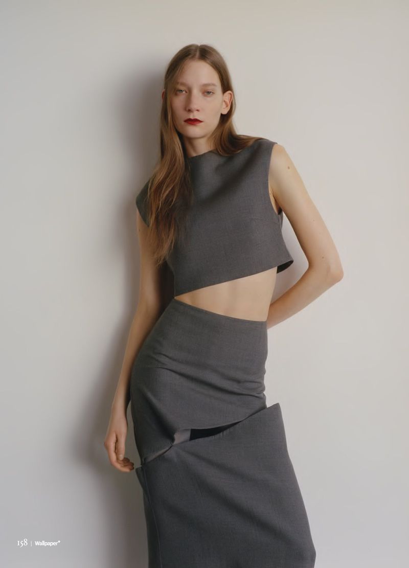 A slender fashion model poses indoors wearing a grey sleeveless top and long skirt against a white wall in a studio-style setting.