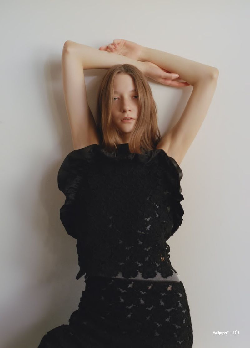 Woman with arms raised poses in a black floral lace top and skirt against a white wall, creating a fashion-forward portrait.