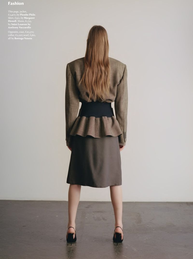 Back view of a fashionable woman with long hair posing in a skirt and jacket, standing against a plain backdrop.