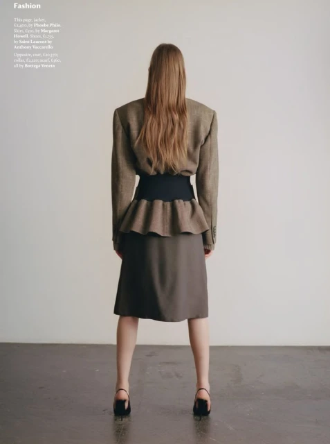 Back view of a fashionable woman with long hair posing in a skirt and jacket, standing against a plain backdrop.
