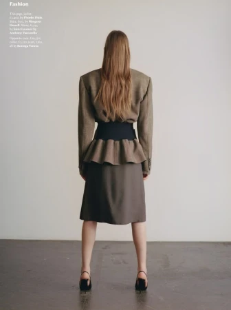 Back view of a fashionable woman with long hair posing in a skirt and jacket, standing against a plain backdrop.