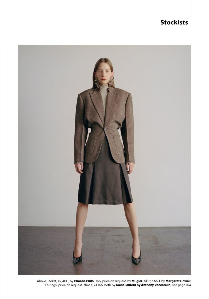 Fashion editorial shot: a woman in a brown blazer, skirt, and black heels poses indoors against a neutral backdrop in a full-length shot.