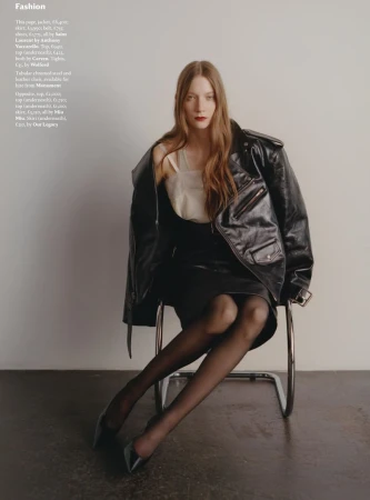 Fashion editorial shot of a poised woman with auburn hair wearing a black leather jacket, black tights, and heels sitting on a chair.