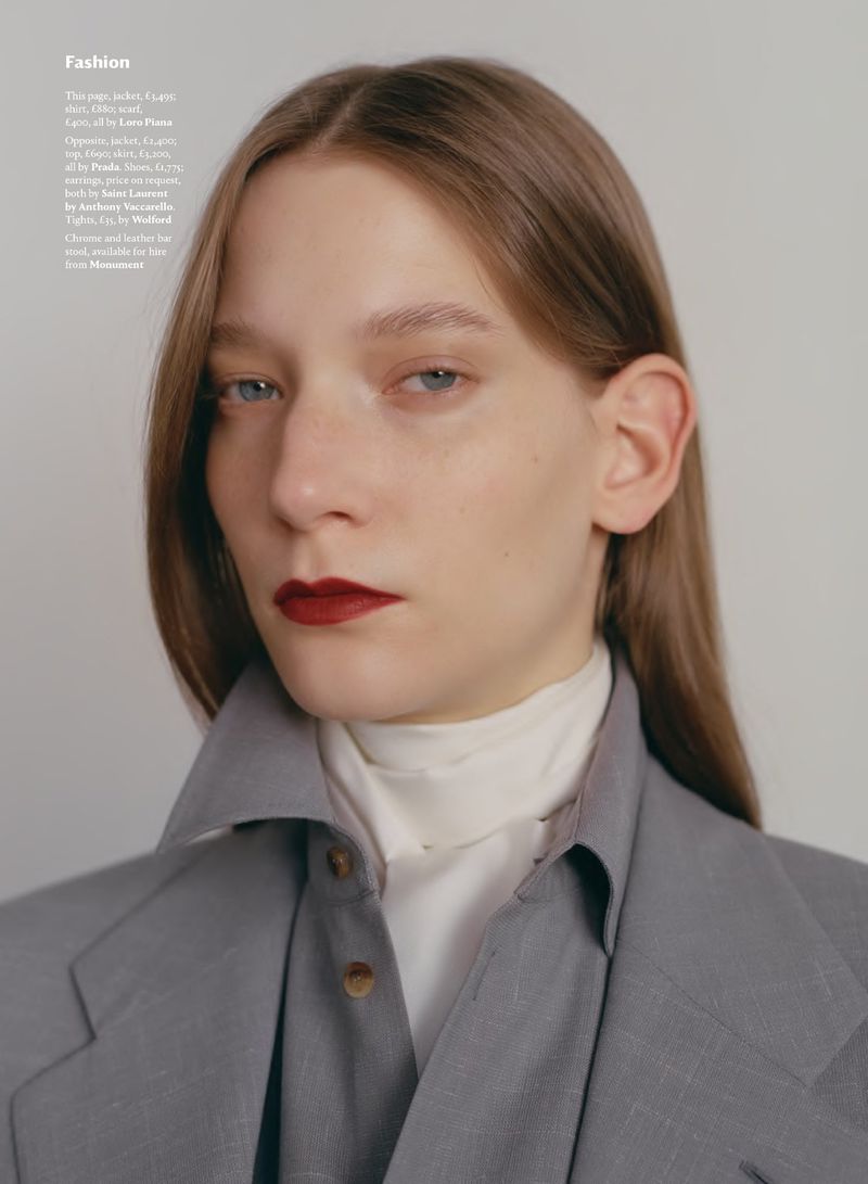 Fashion editorial shot of a young adult woman with reddish lipstick and straight light brown hair, wearing a grey suit jacket and collared shirt.