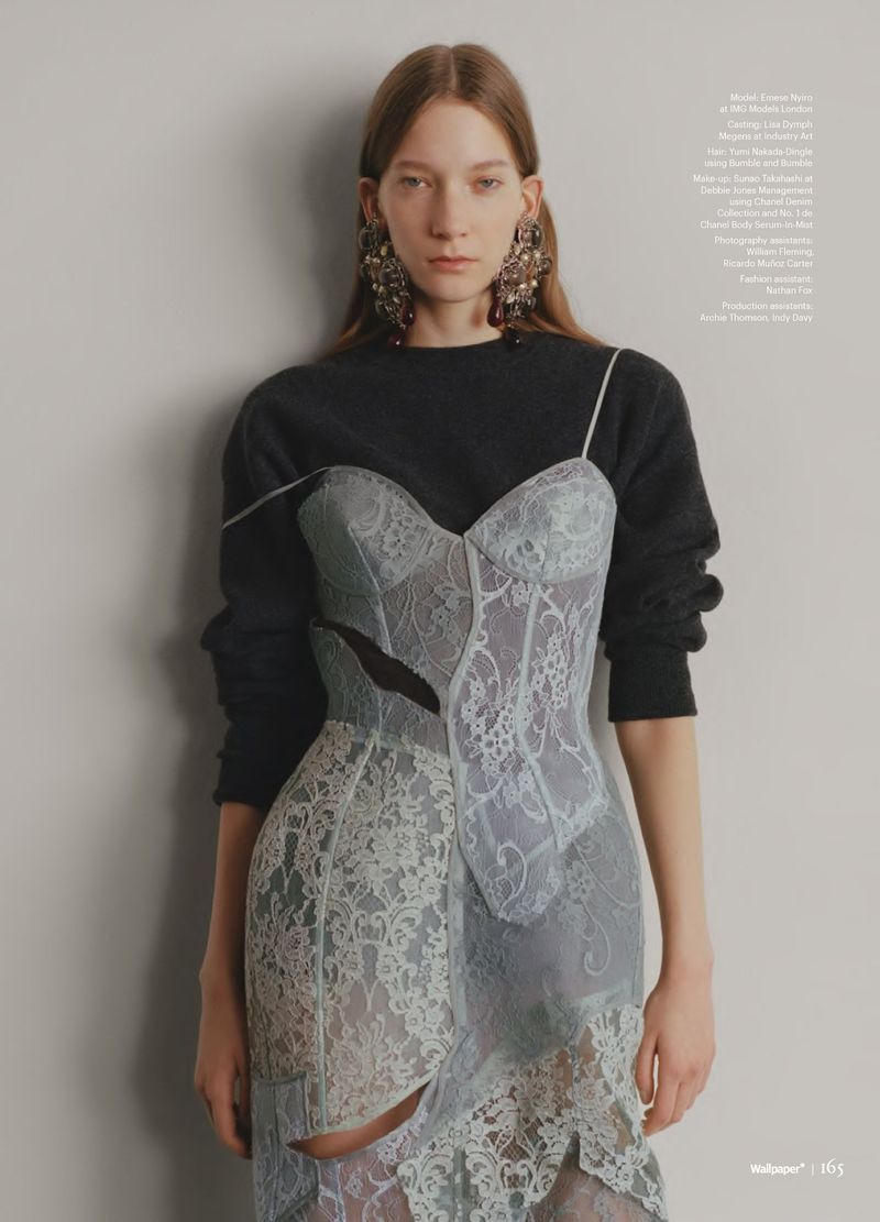 Editorial fashion shot of a woman wearing a light blue lace dress with a black long-sleeved shirt, large statement earrings, indoors against a grey wall.