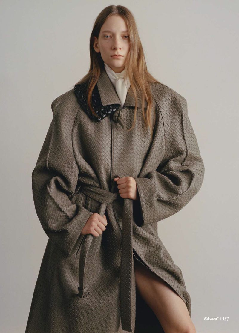 A young woman poses in a brown patterned woven trench coat with a tie belt in front of a solid light background in a studio setting.