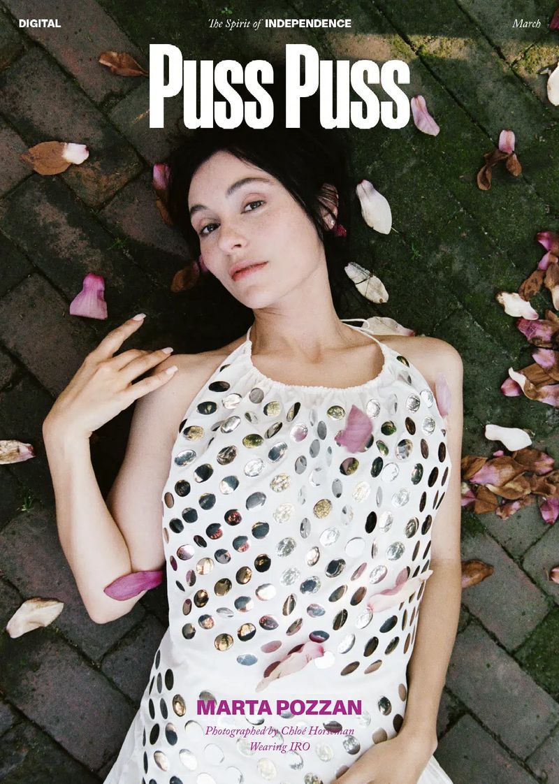Magazine cover featuring a model with dark hair lying on a brick surface surrounded by flower petals. Wearing an embellished white dress.