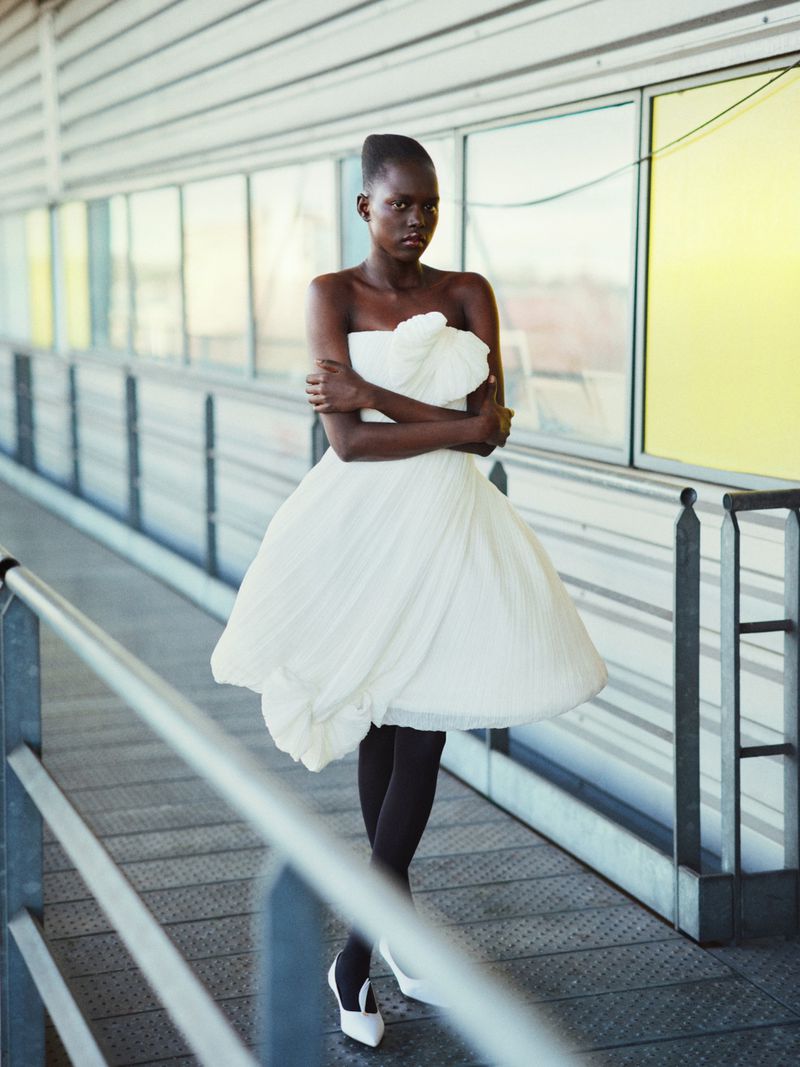 Elegant woman poses in flowing white dress and heels on an elevated walkway with metal railings and colorful window accents.
