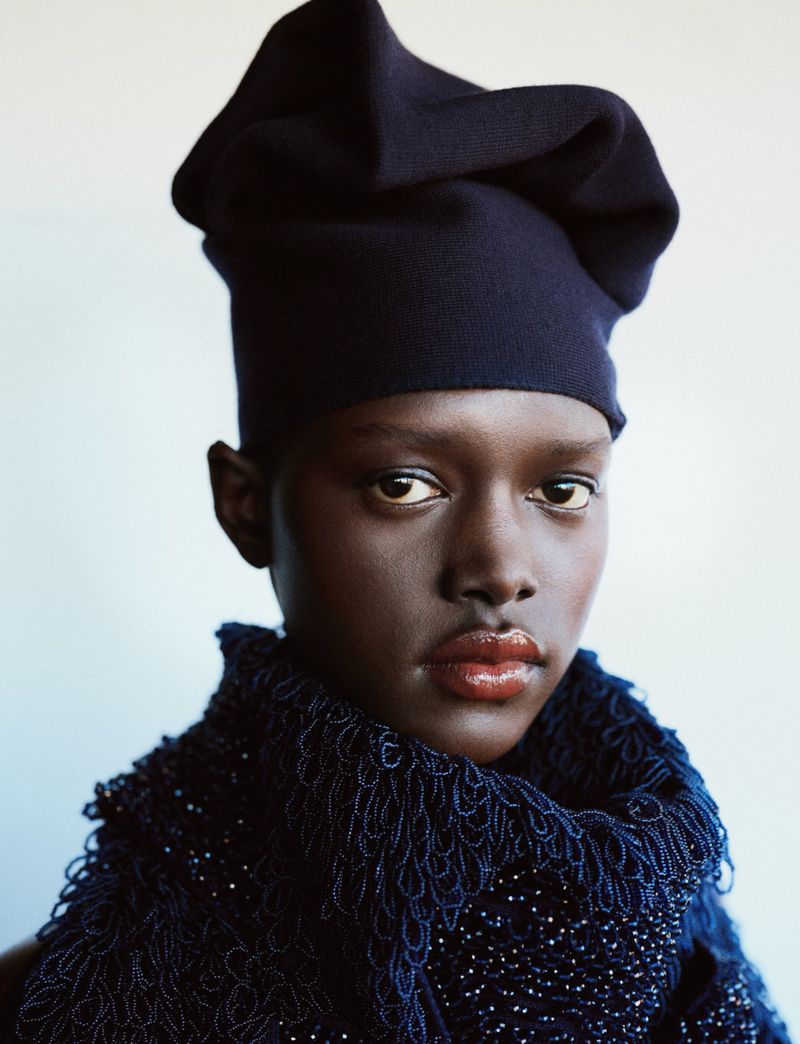 Close-up studio portrait of an adult Black woman wearing a dark blue knit hat and a beaded fringed scarf against a gradient background.