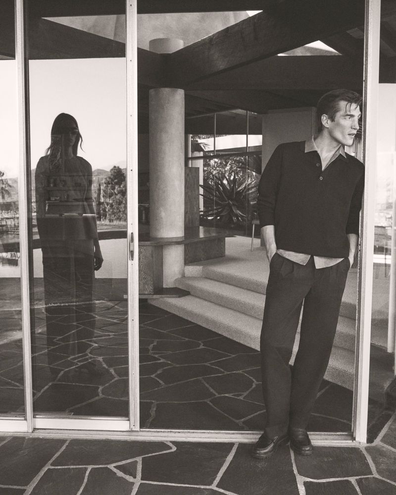A man in a polo shirt leans against a doorway in a modern residence, while a woman's reflection is visible in the adjacent glass door.