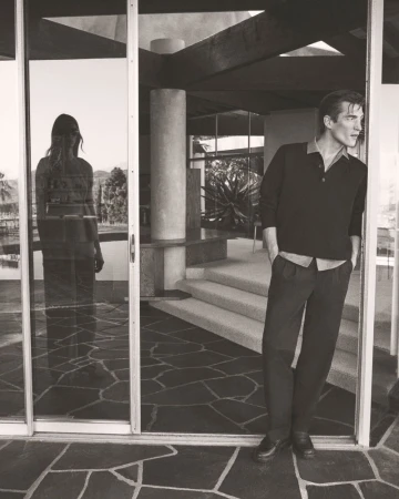 A man in a polo shirt leans against a doorway in a modern residence, while a woman's reflection is visible in the adjacent glass door.