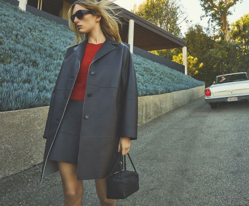 A blonde-haired woman in a black coat and sunglasses stands on a driveway next to a retro car and modern house with a drought-tolerant garden.