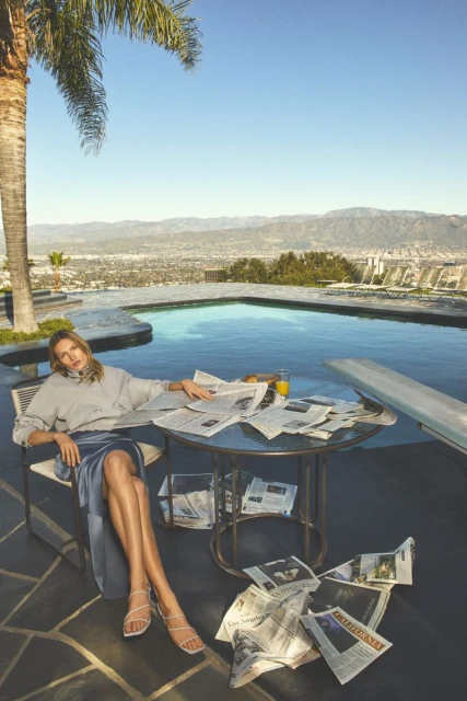 Adult female relaxes outdoors near a pool, dressed in a sweatshirt and skirt, surrounded by newspapers on a sunny day.