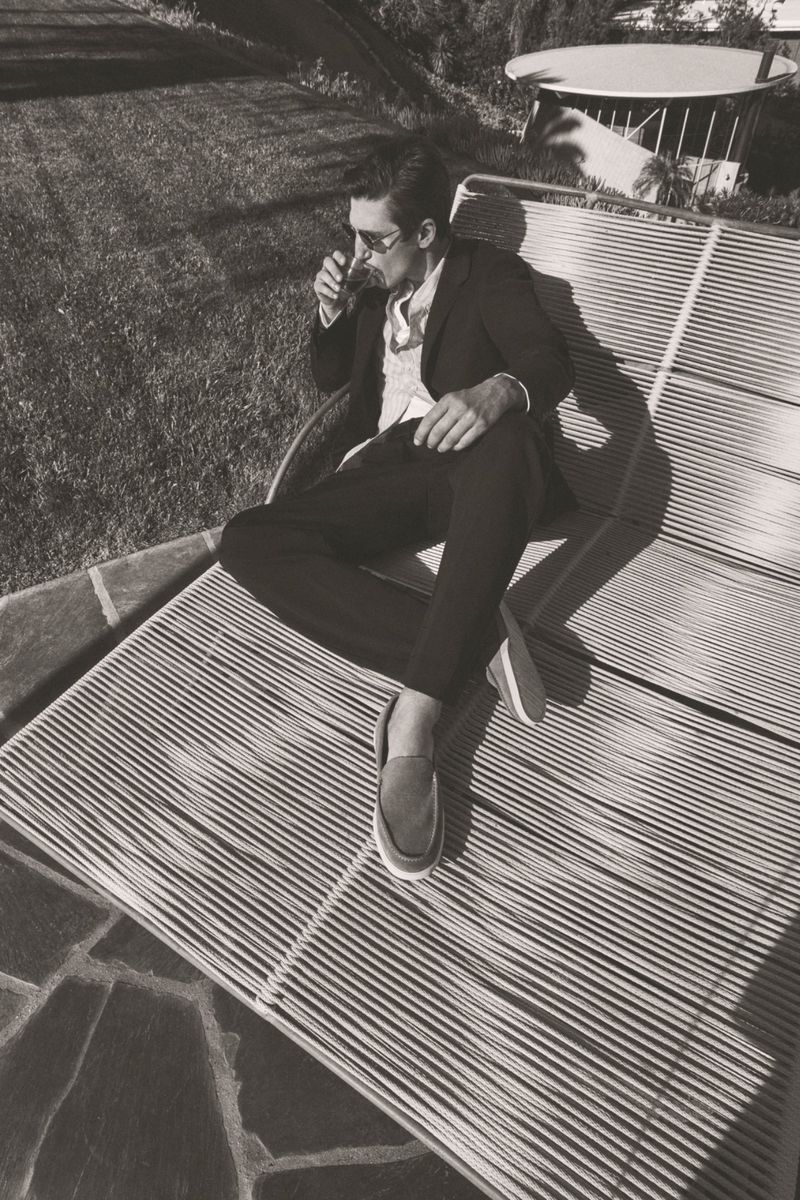 Black and white shot of a man in a suit reclining on a striped lounge chair outdoors, drinking a beverage in the sunlight.
