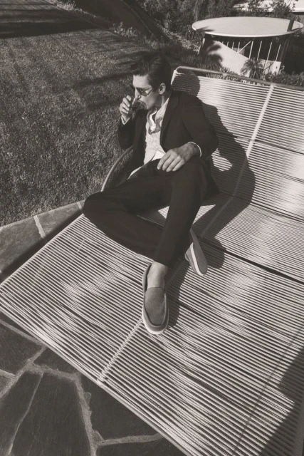 Black and white shot of a man in a suit reclining on a striped lounge chair outdoors, drinking a beverage in the sunlight.
