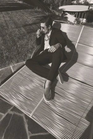 Man wearing a dark suit and loafers reclines on a slatted outdoor lounger, drinking from a glass in a bright sunny garden setting.