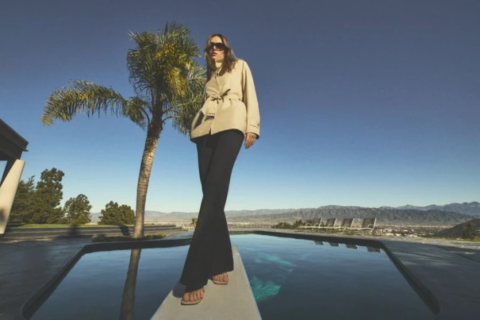Adult woman standing on ledge over infinity pool wearing a jacket, sunglasses, and flared black pants against the blue sky.