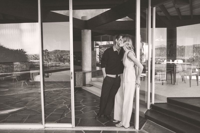 Monochrome shot of a couple kissing indoors, with a modern home interior visible through sliding glass doors and mountain views.