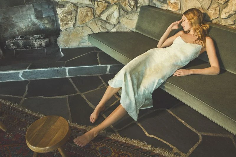 Woman in a white dress reclines on a green couch, near a stone fireplace, and rests her hand on her forehead in a contemplative pose.