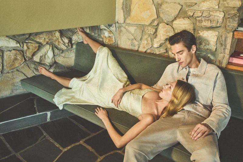 A couple rests together on a green couch. The woman is lying down with her head on the man's lap in a bright indoor setting.