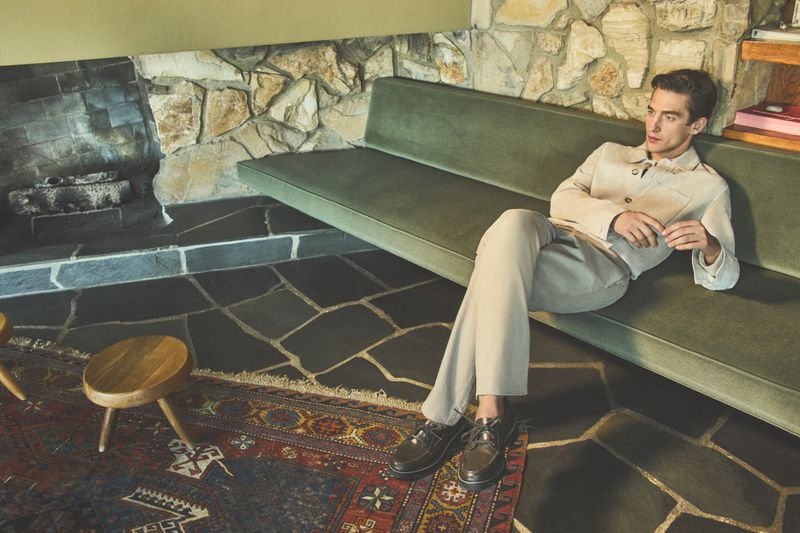 Adult male model wearing beige jacket and pants lying on a green sofa indoors with stone fireplace, patterned rug, and wooden stool.