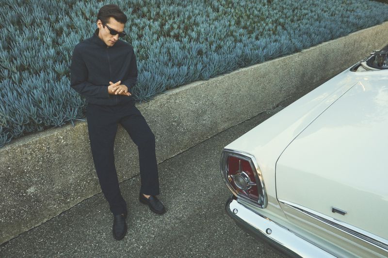 Man wearing dark jacket, trousers, and sunglasses leans against a stone wall with succulents, next to a vintage white convertible car.