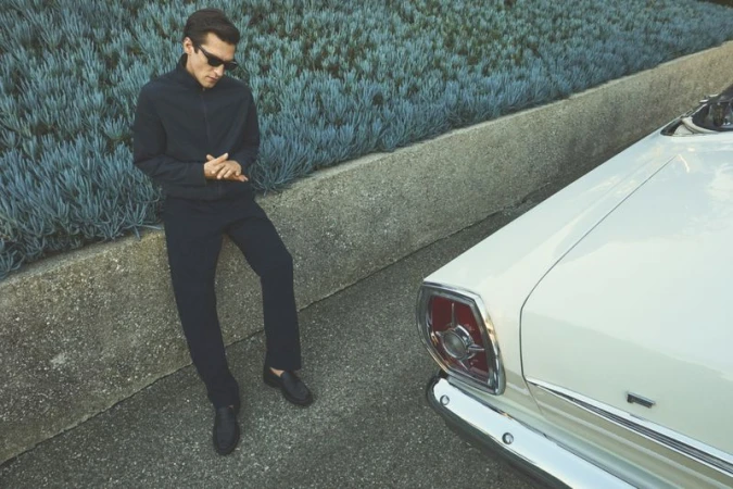 Man wearing dark jacket, trousers, and sunglasses leans against a stone wall with succulents, next to a vintage white convertible car.